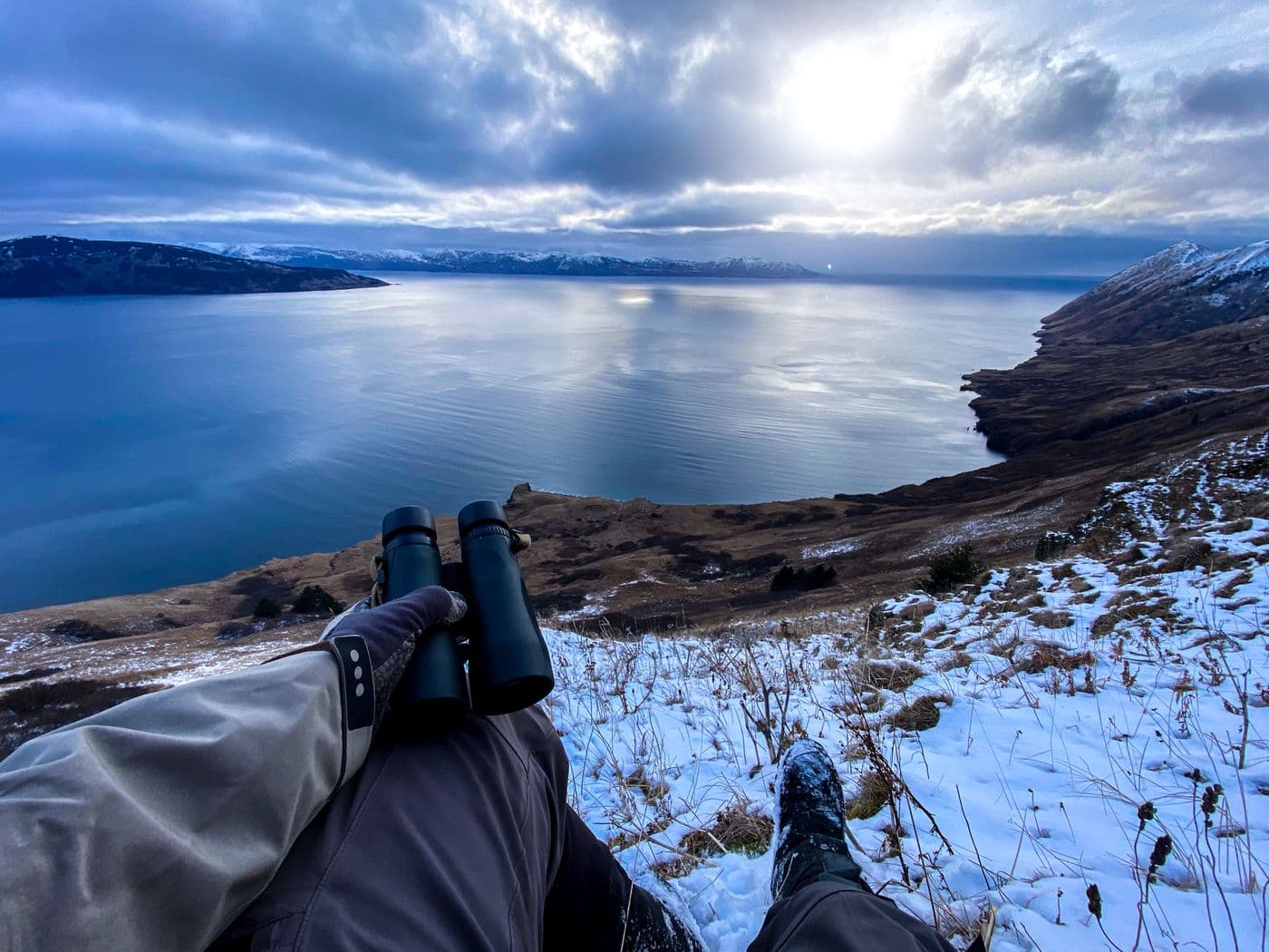 A gloved hand steadying binoculars above open water on a snowy slope at dusk.