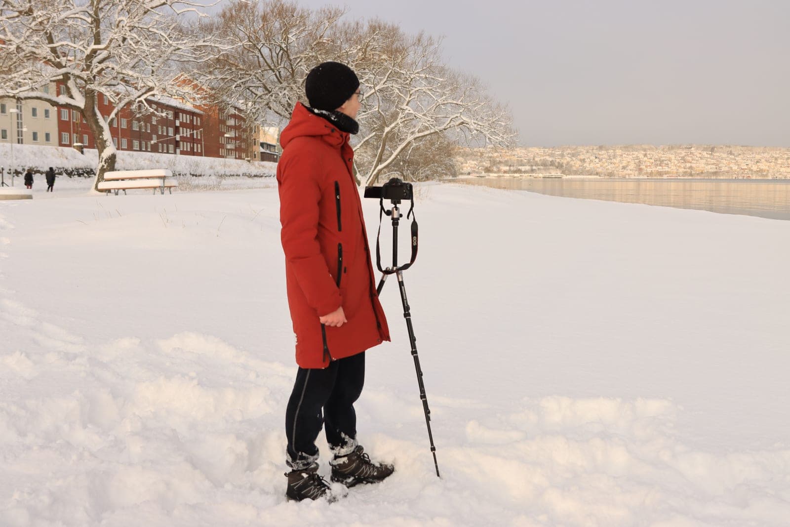 A person in a heavy winter coat standing in deep snow beside a tripod with its legs planted in the snow, looking out over open water.